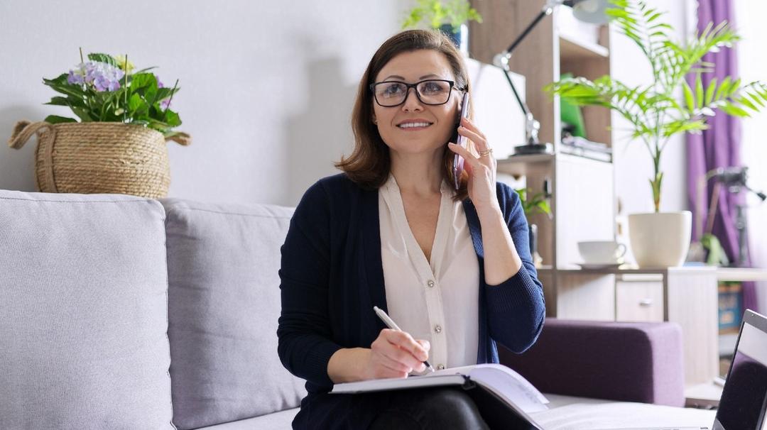 Mature woman working remotely at home, business female freelancer sitting on sofa with laptop, talking on phone, taking notes in business notebook.