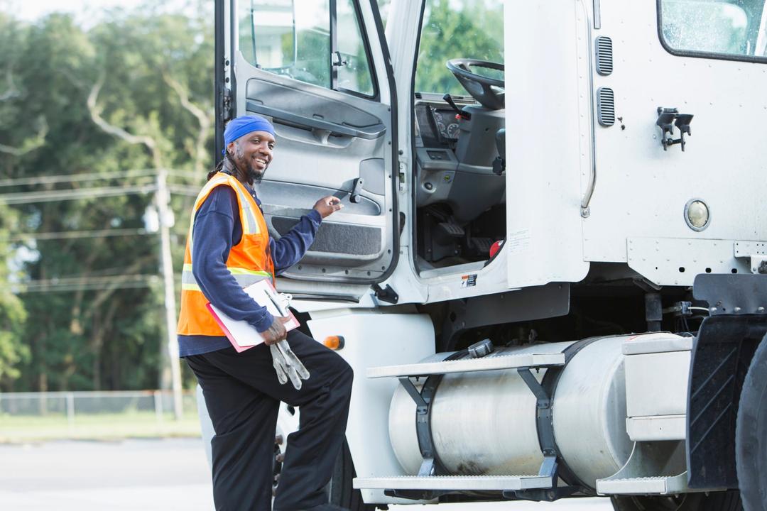 A semi-truck driver climbing into the truck cab, smiling at the camera. The mature black man in his 40s is wearing a reflective vest.