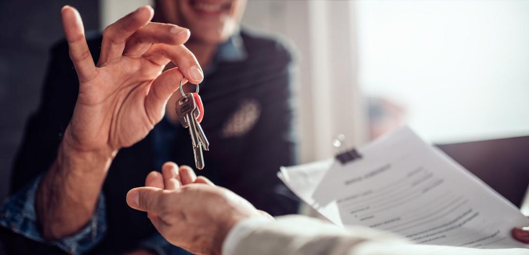 A person receives keys from another, smiling, with a document in the background, indicating a possible transaction or agreement.