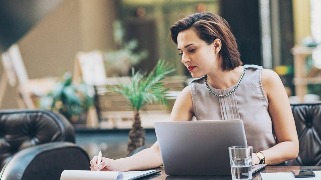 A professional woman working on a laptop in a stylish office space, taking notes in a notebook, with a potted plant in the background.