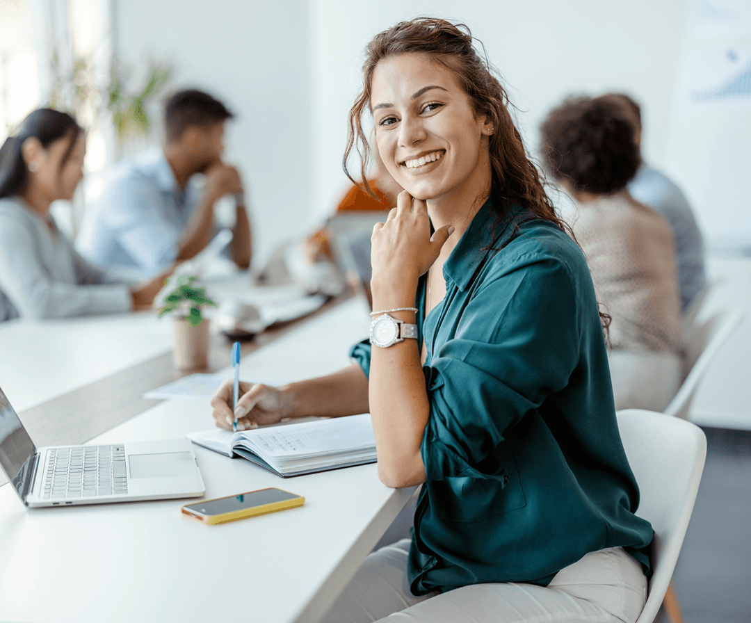 A smiling young woman in a teal blouse working at a laptop in a modern office with colleagues in the background.