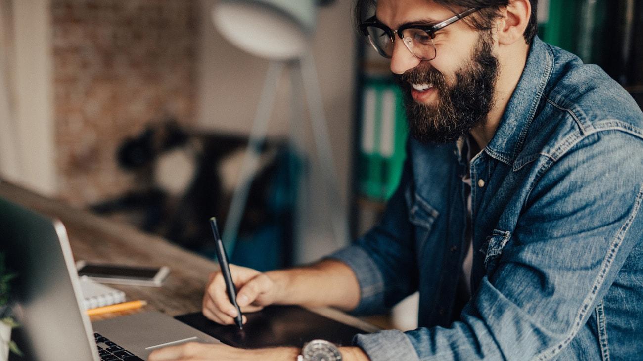 A smiling man with a beard, wearing glasses and a denim shirt, uses a graphics tablet and laptop in a modern office.