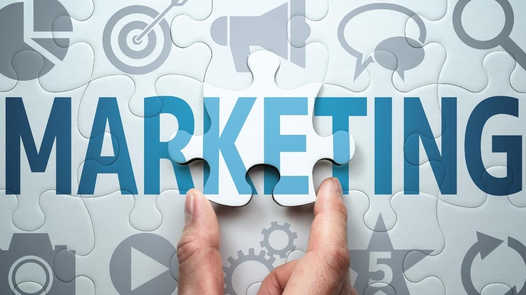 Two hands placing the final piece of a puzzle to complete the word "marketing" surrounded by various business icons.