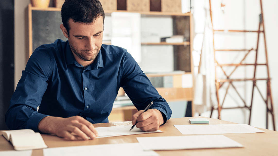 A man in a blue shirt concentrating while writing on a paper at a wooden desk, with books and an eraser nearby, in a modern office setting.