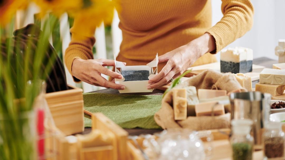 A person in a yellow sweater packaging a handmade soap bar at a craft table surrounded by various natural ingredients and materials.