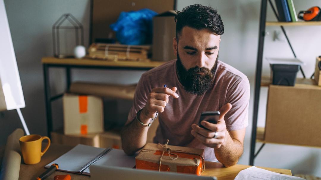 Man with beard using smartphone at desk with notebook, wrapped parcel, and coffee mug, in a home office setting.