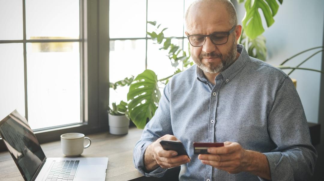 A balding middle-aged man with glasses, using a smartphone and holding a credit card, seated at a desk with a laptop and coffee cup nearby.