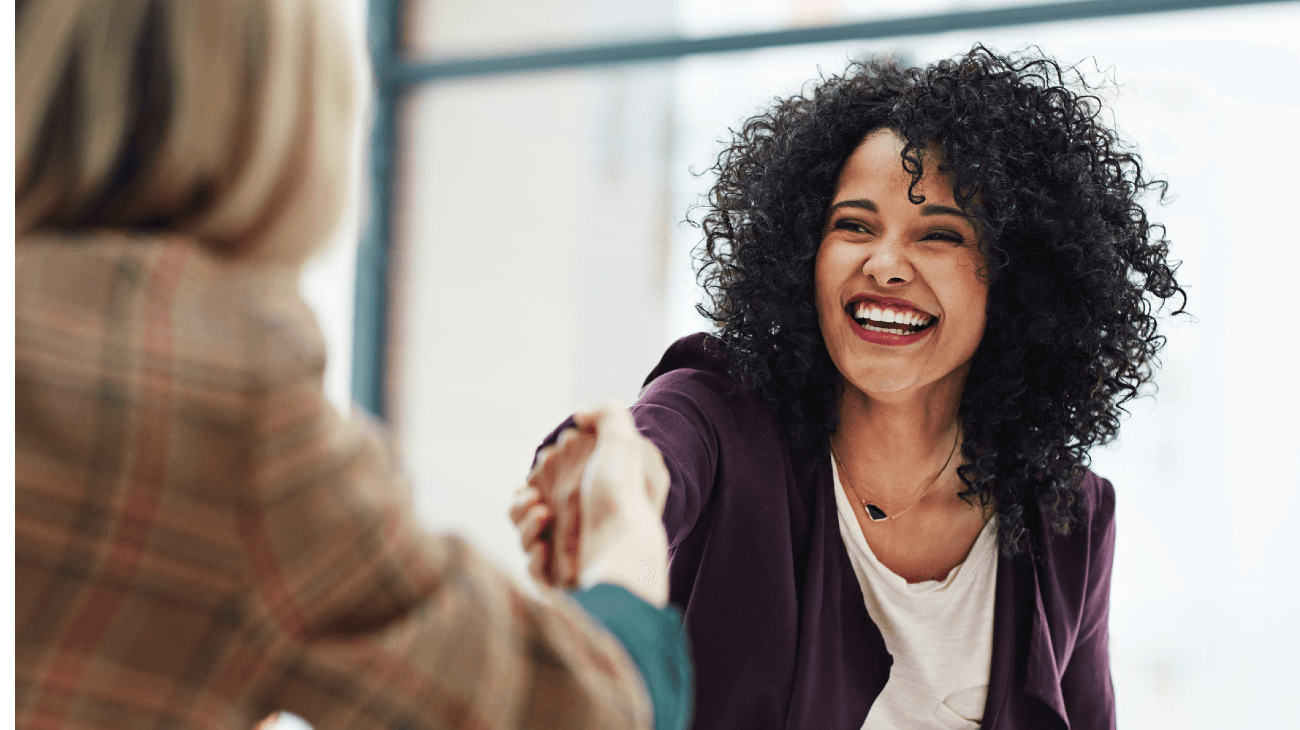 Two women in an office setting, smiling and shaking hands, one with curly hair and a purple blazer, the other partially visible.