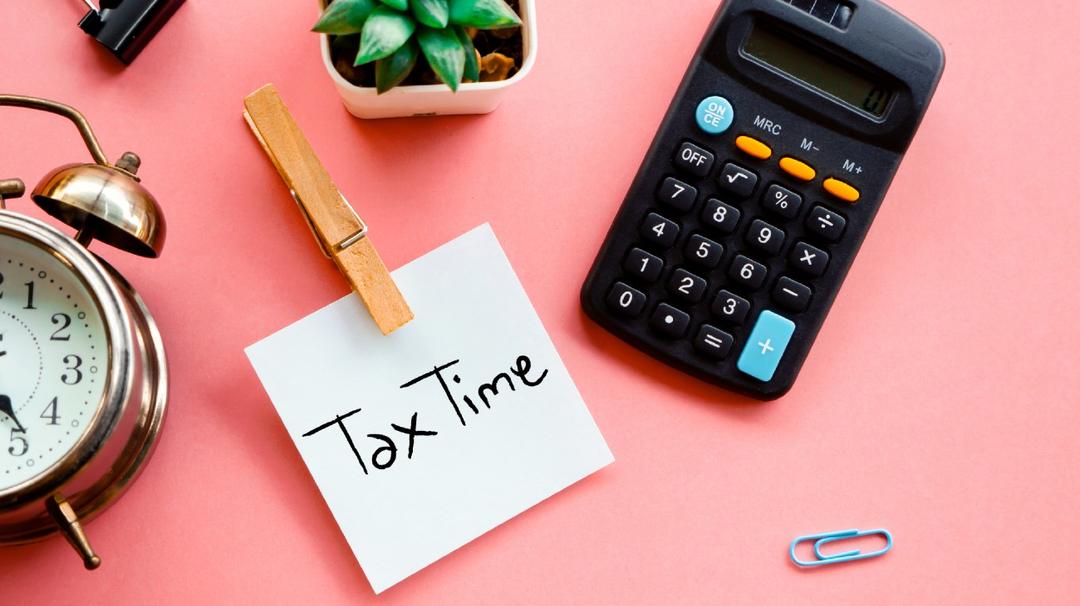 A desktop with a calculator, a clock, a potted plant, and a note saying "tax time" on a pink background.
