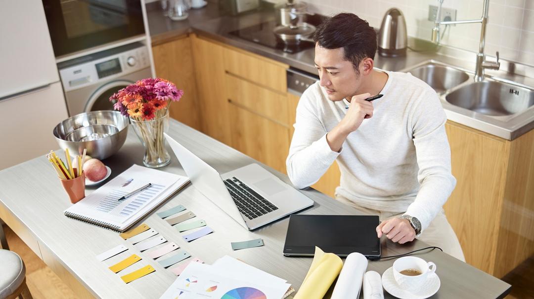 Man at kitchen counter with laptop and documents spread out in front of him