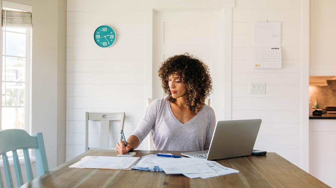 A woman with curly hair works at a home office desk, writing notes beside a laptop in a well-lit room with white walls and a blue clock.